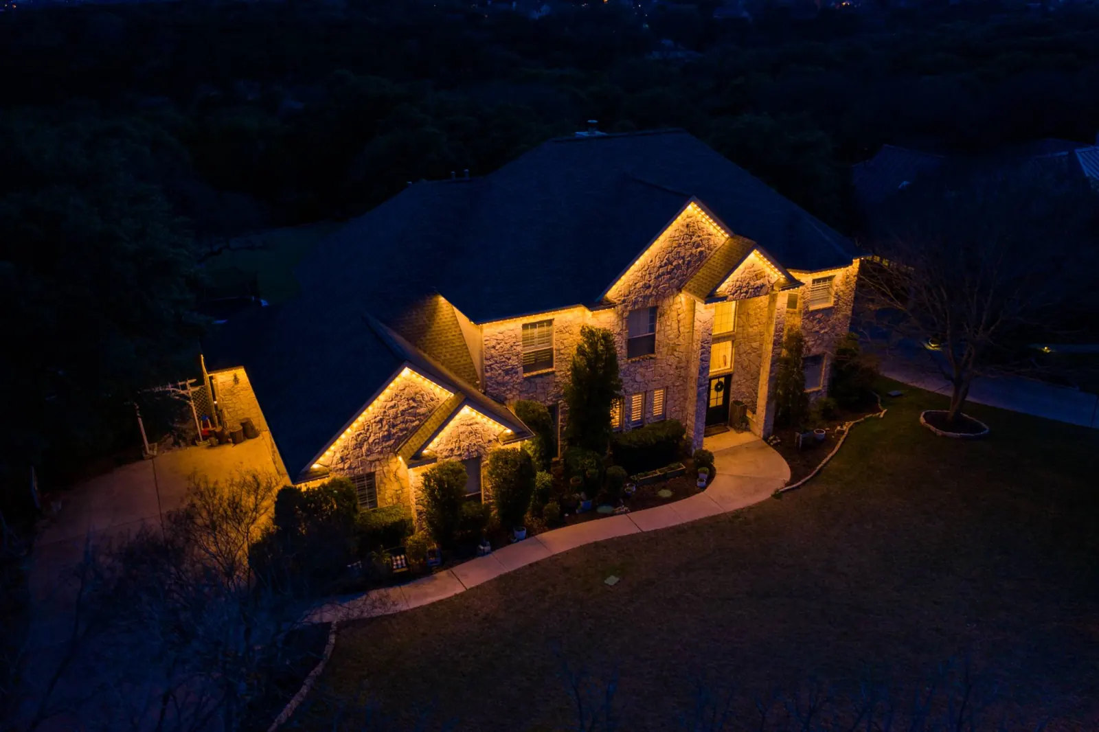 Aerial view at dusk of a large stone house with warm roofline lighting and uplit trees along the drive