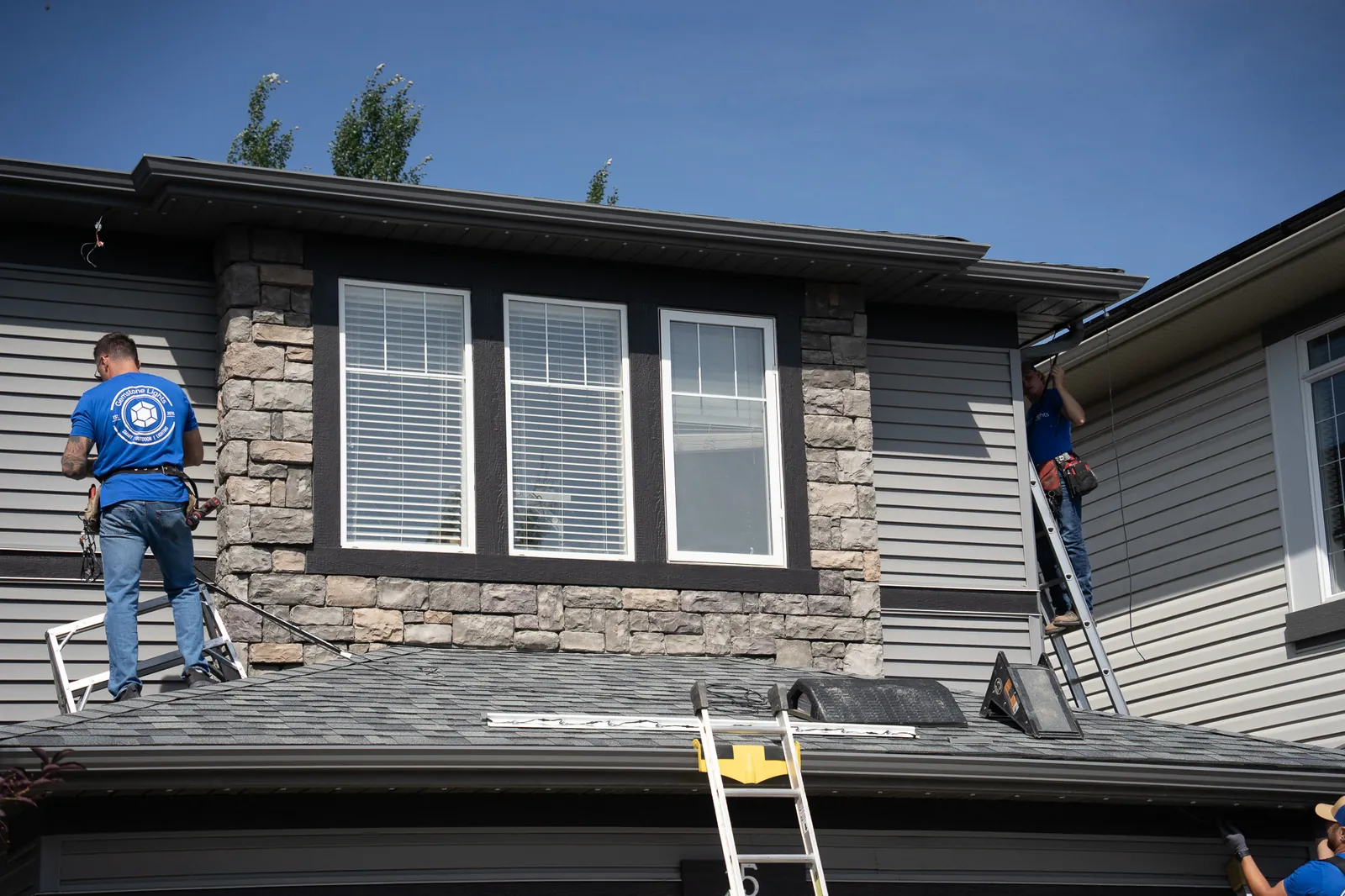 Two installers fitting permanent track lights under the roofline of a stone and siding home