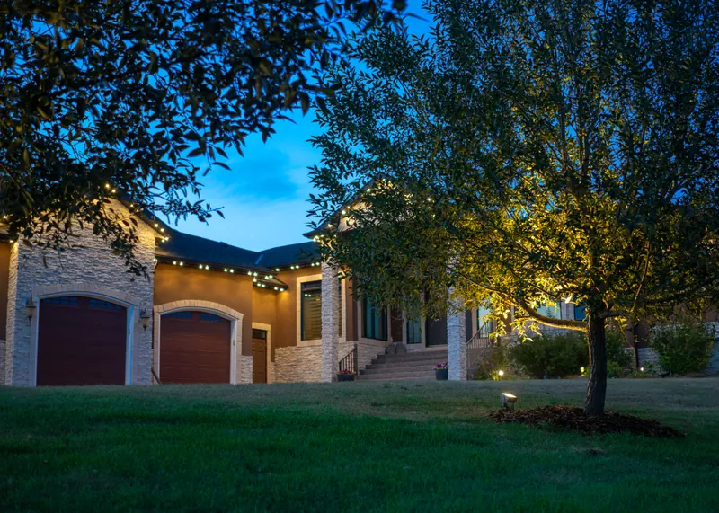 Stone house at dusk with warm white roofline lights and a young tree on the lawn lit from below by a ground light
