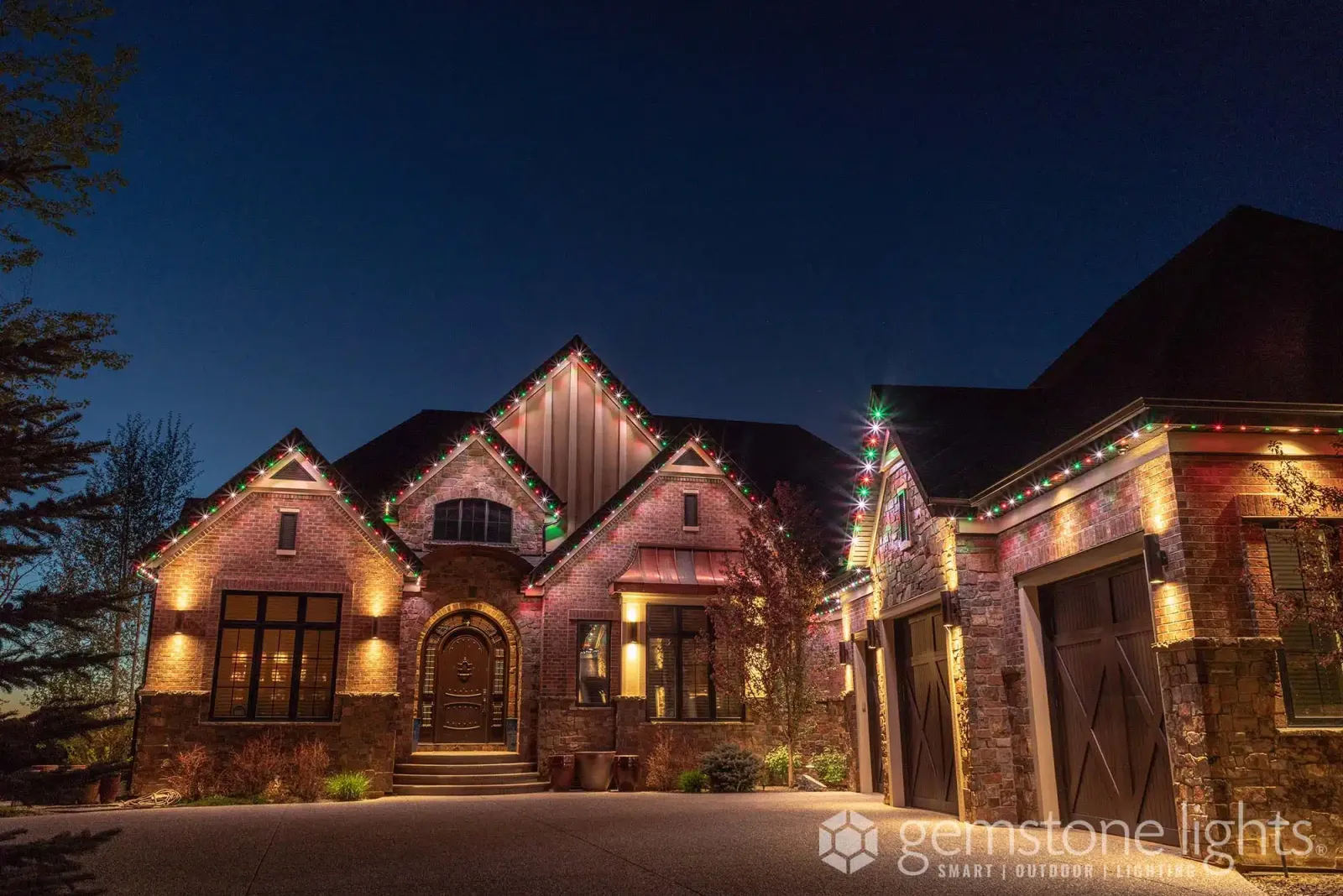 Stone and brick home with several steep gables, each outlined in warm white lights under a twilight sky