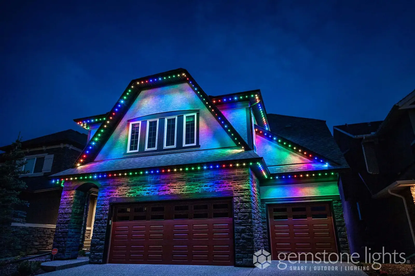 Two-story home with a steep front gable lit in a rainbow sequence from the roof peak down to the garage