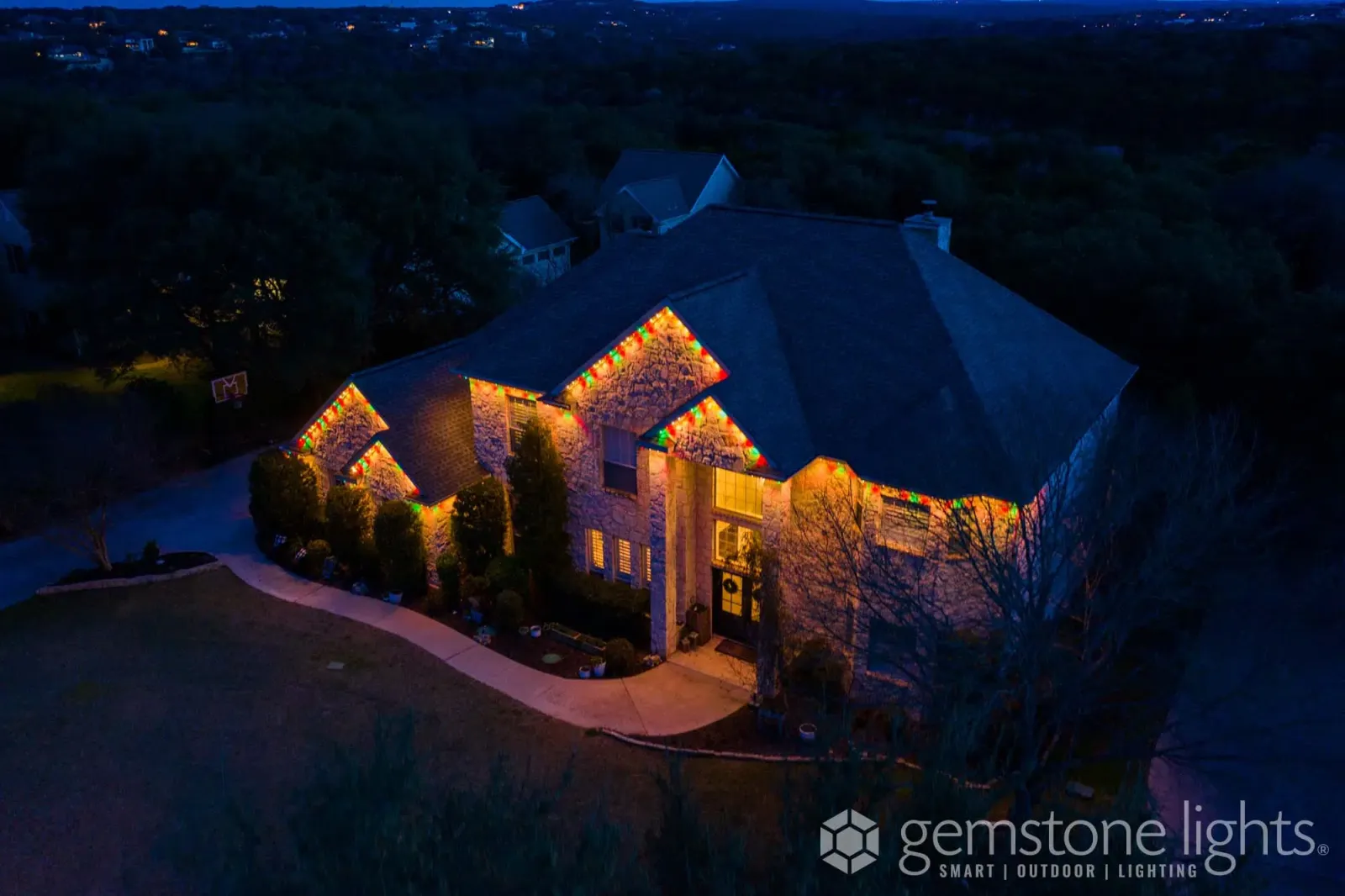 Aerial night view of a large home on a wooded lot, its rooflines traced in warm white lights