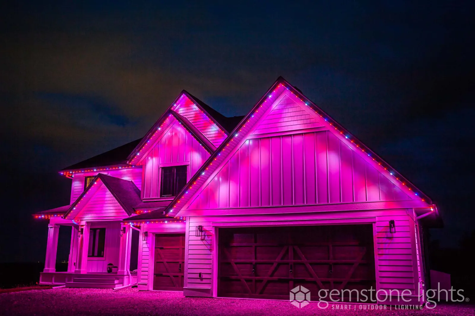 Farmhouse-style home with board-and-batten gables glowing magenta and pink at night